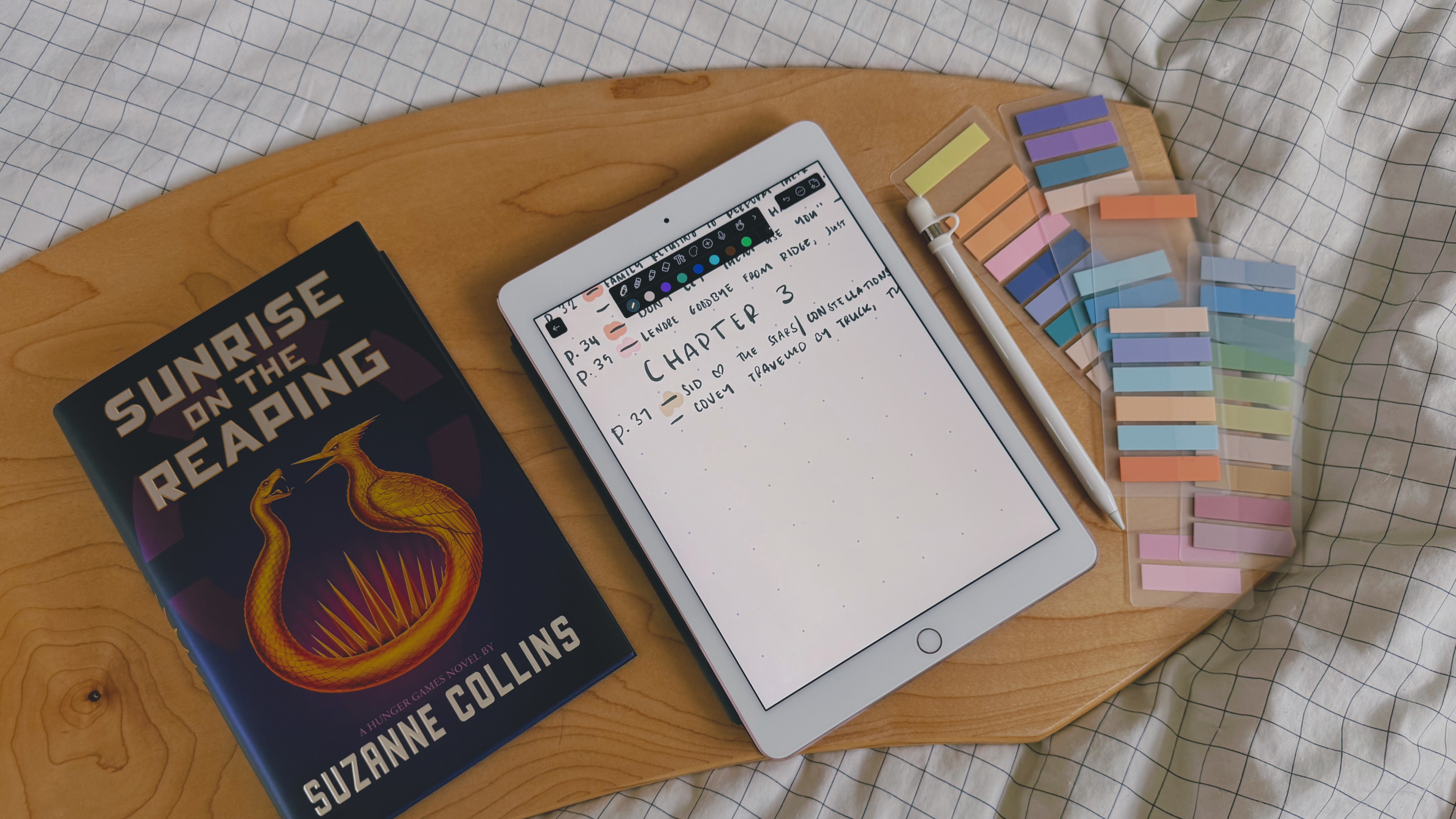 Photo of a wooden surface on a white bedsheeet. On the wooden surface is a copy of Sunrise on the Reaping by Suzanne Collins, an iPad with a notes app open, an Apple pencil, and sticky book tabs.