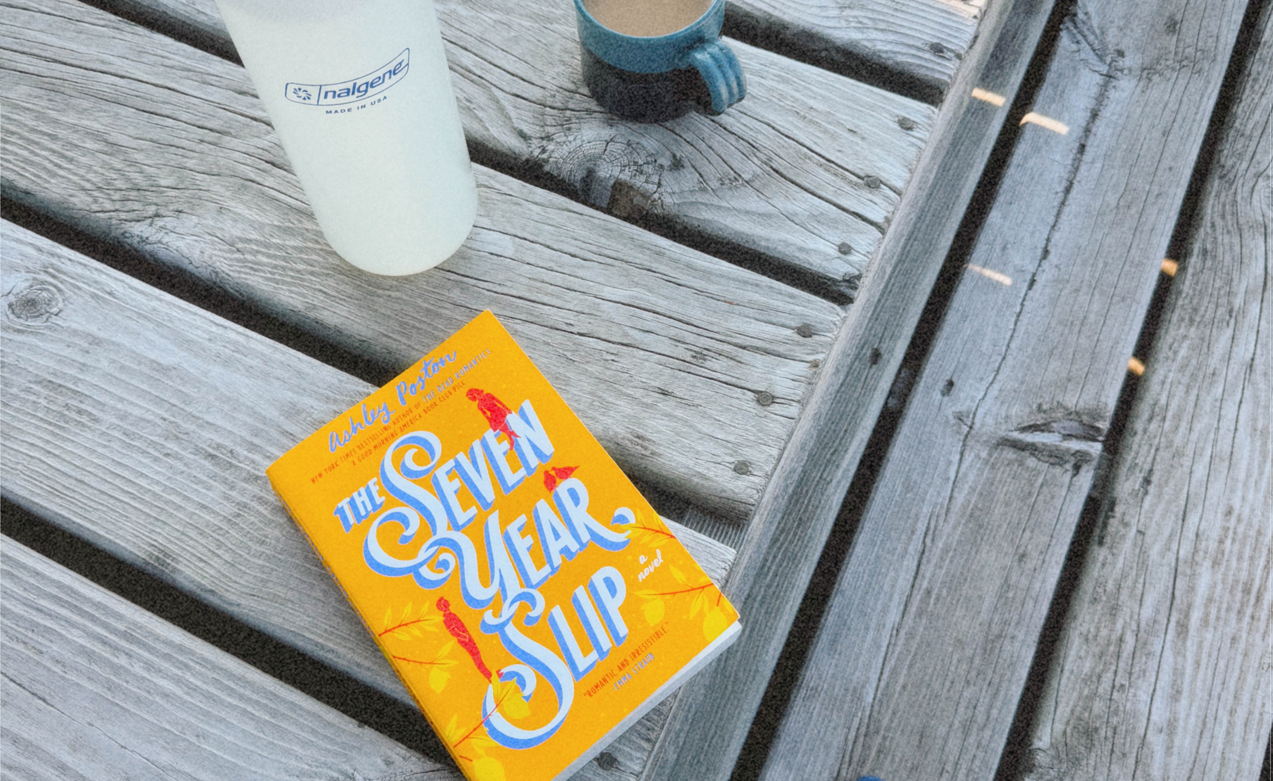 Photo of a worn wooden deck with a bright yellow book, The Seven Year Slip by Ashley Poston, resting on it next to a white nalgene water bottle and a ceramic mug of coffee.