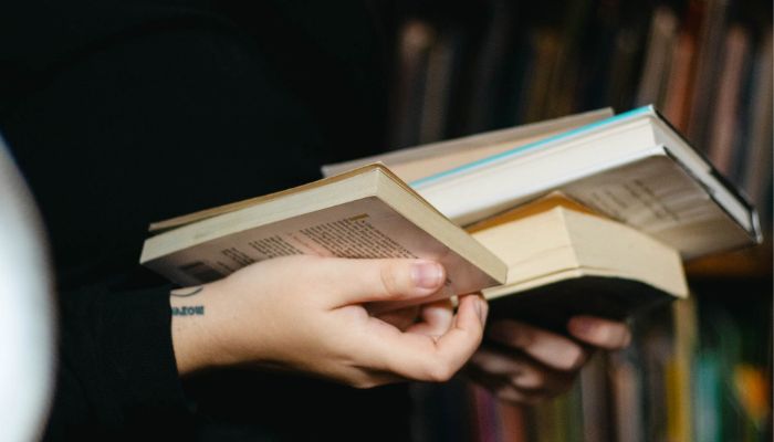 Close up photo of someone's hands holding a few books. There are bookshelves in the background.