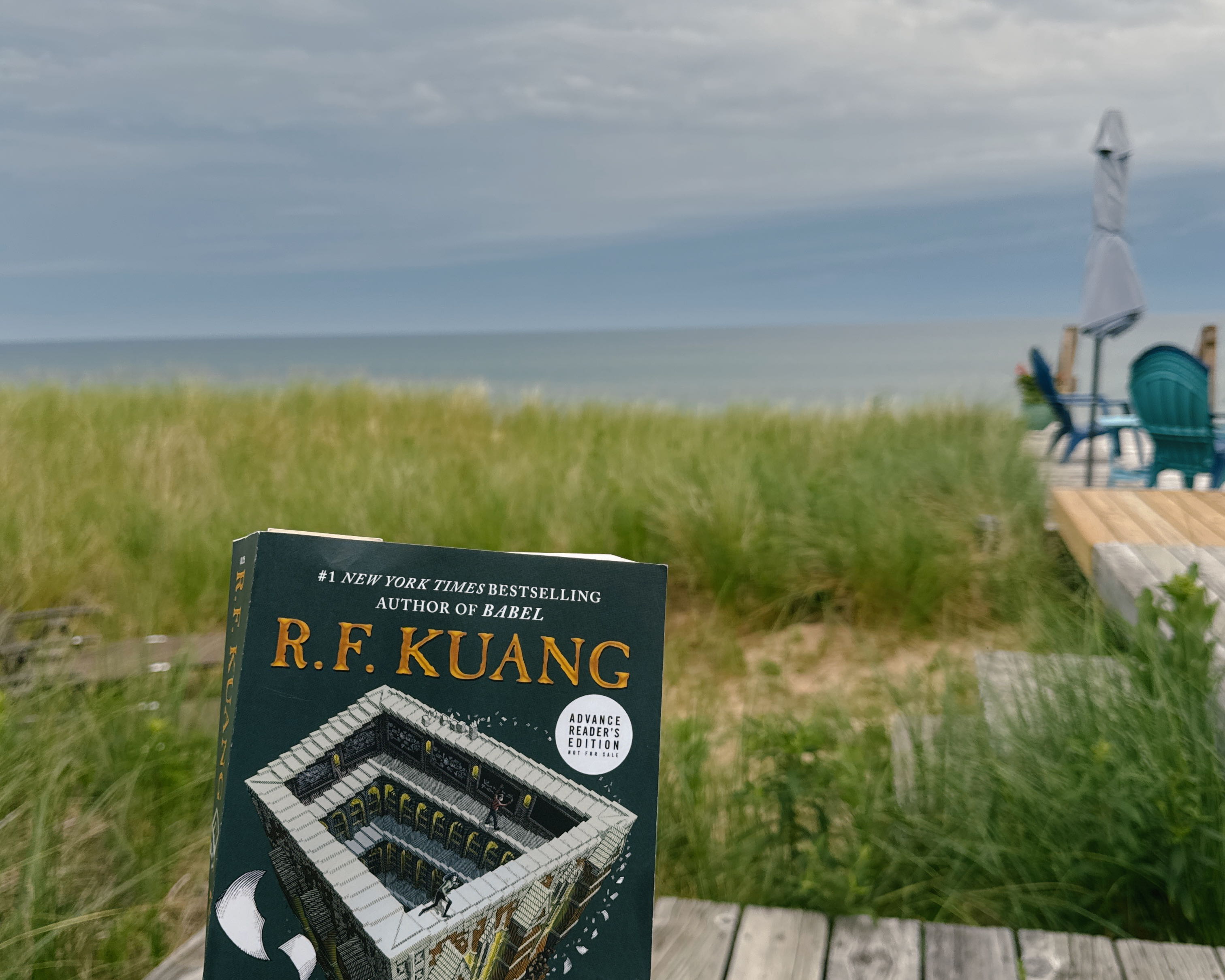 Photo of a book in front of grassy dunes and a lake in the distance. The book cover is dark green and has a contunuious staircase art. The top of the book shows the author name, R.F. Kuang.