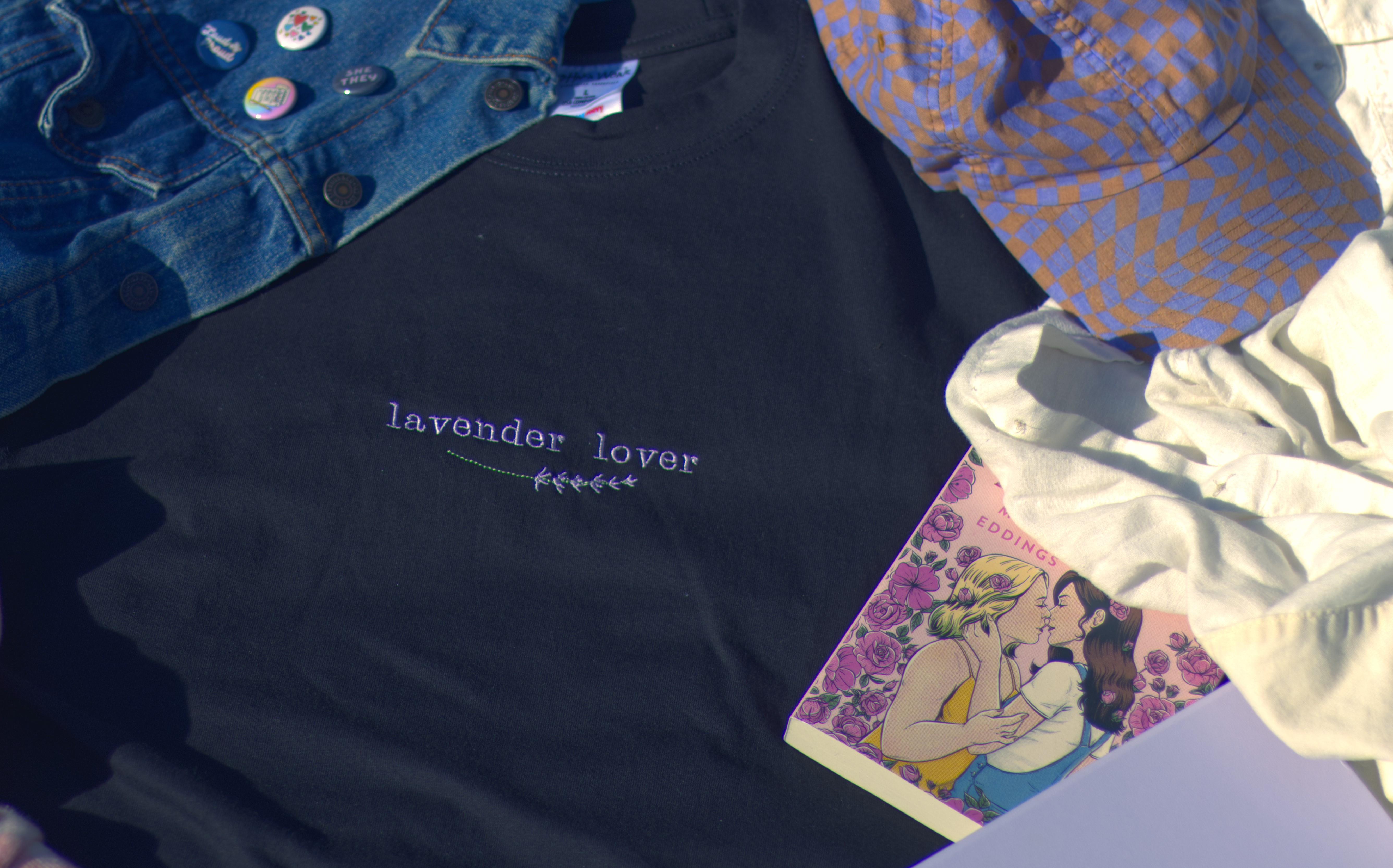 Photo looking down at a black t-shirt that says lavender lover in purple embroudery with a sprig of lavender. Next to the shirt there is the book Late Bloomer which features a cover with two women kissing surrounded by flowers.