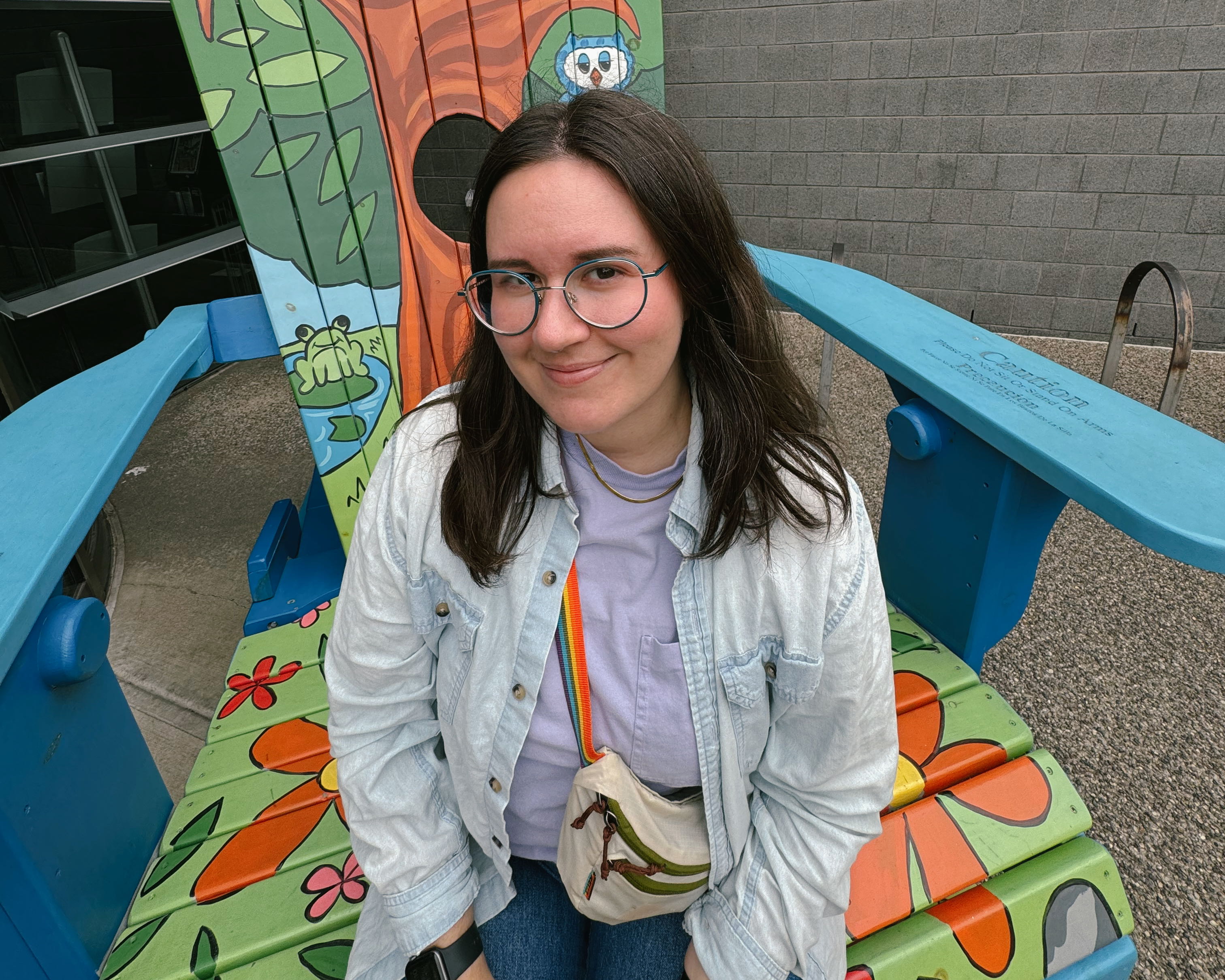 Photo of Bella sitting in an oversized lawn chair. The chair is brightly painted in blue and green and orange. Bella is smiling and looking up at the camera.