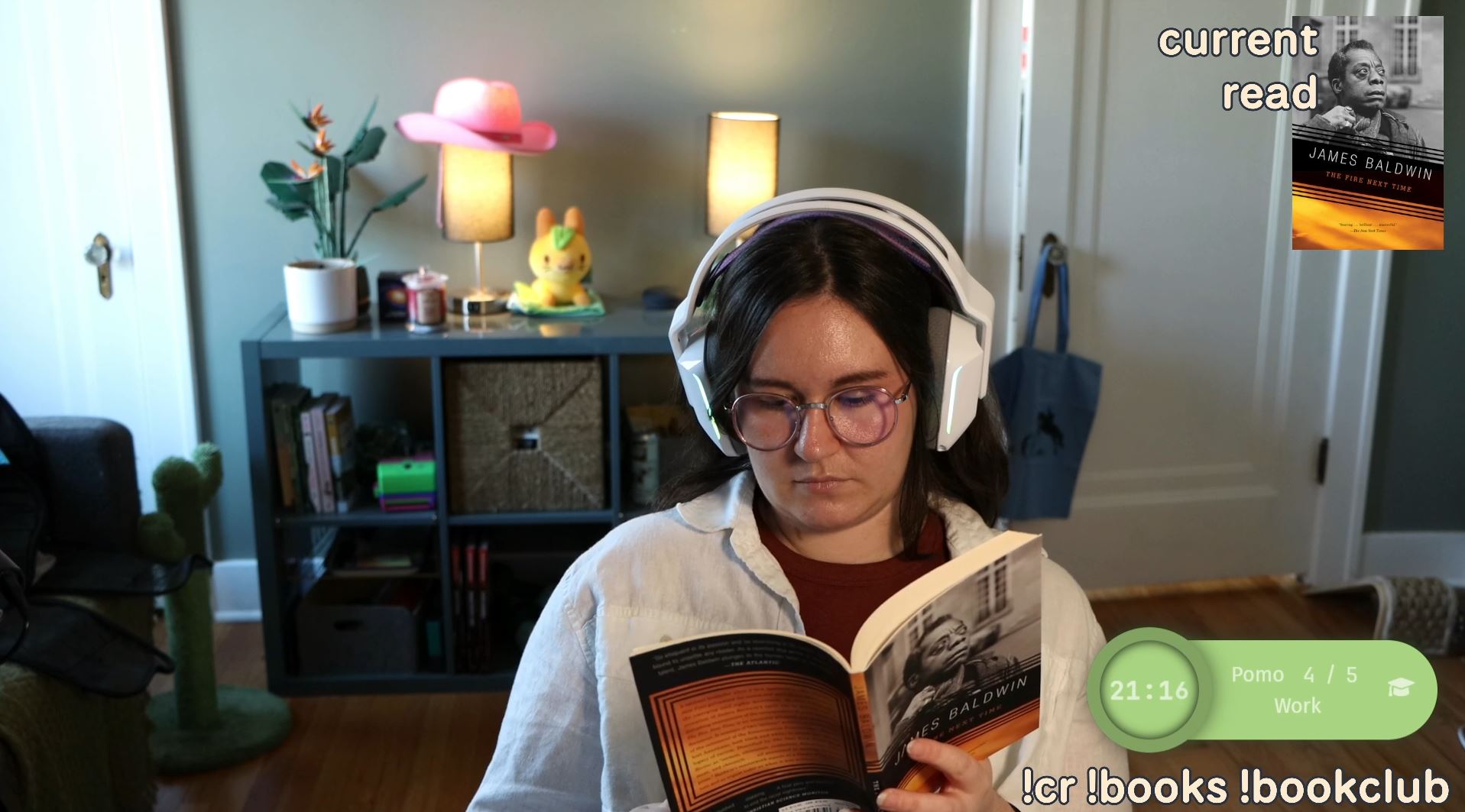 Photo in Bella's streaming room of Bella reading a book during stream. Bella is looking down and reading The Fire Next Time by James Baldwin. There is a green pomodoro task timer on the bottom of the screen and an image of the book cover with the words current read at the top.