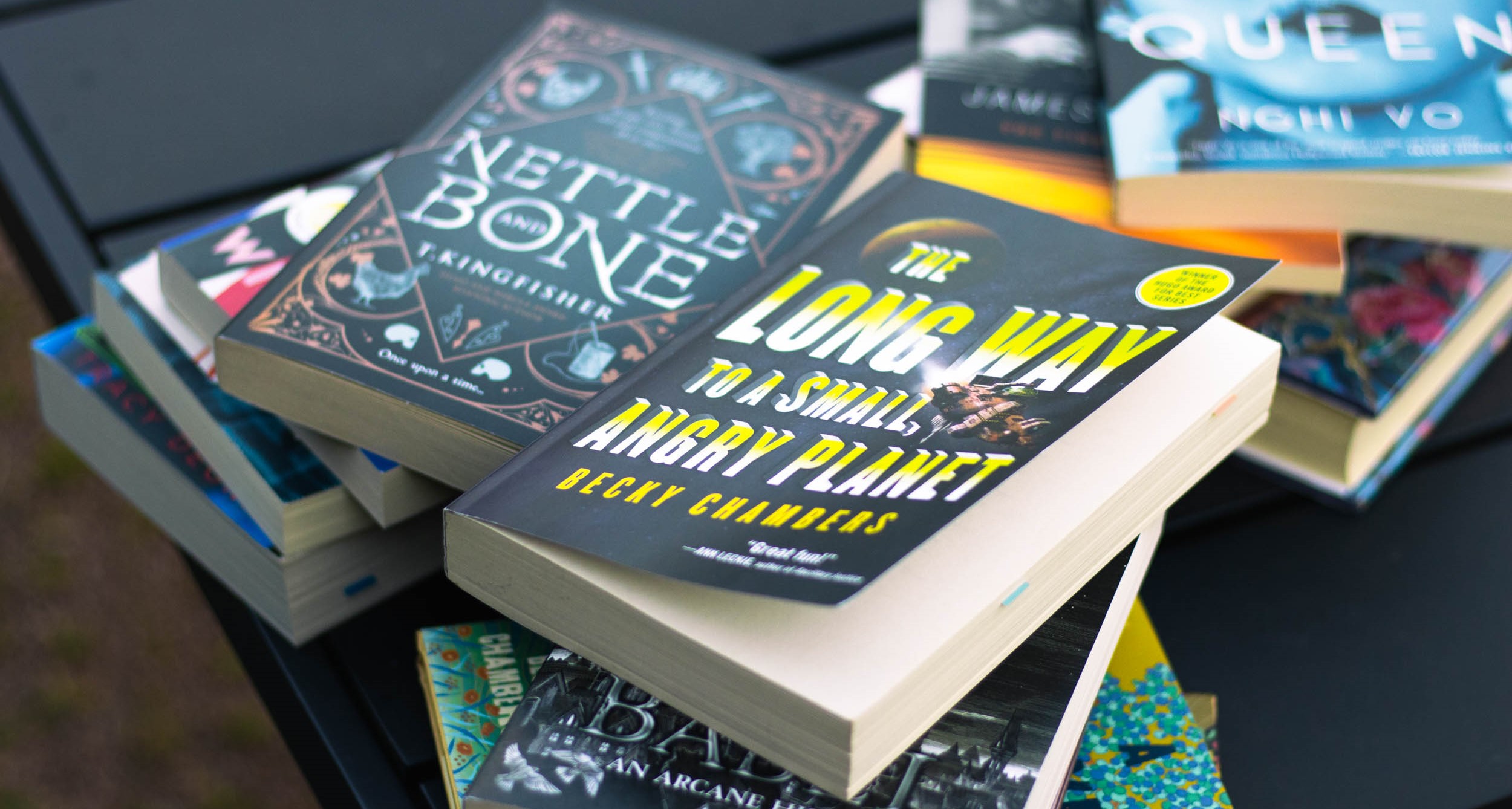 Several books stacked in piles on a black table. Books on top of the piles that are visible include Nettle and Bone by T. Kingfisher and The Long Way To A Small, Angry Planet by Becky Chambers.