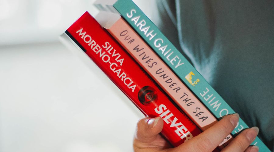 Photo of Bella holding three books in their arm. The books shown are The Echo Wife by Sarah Gailey, Our Wives Under the Sea by Julia Armfield, and Silver Nitrate by Silvia Moreno-Garcia.