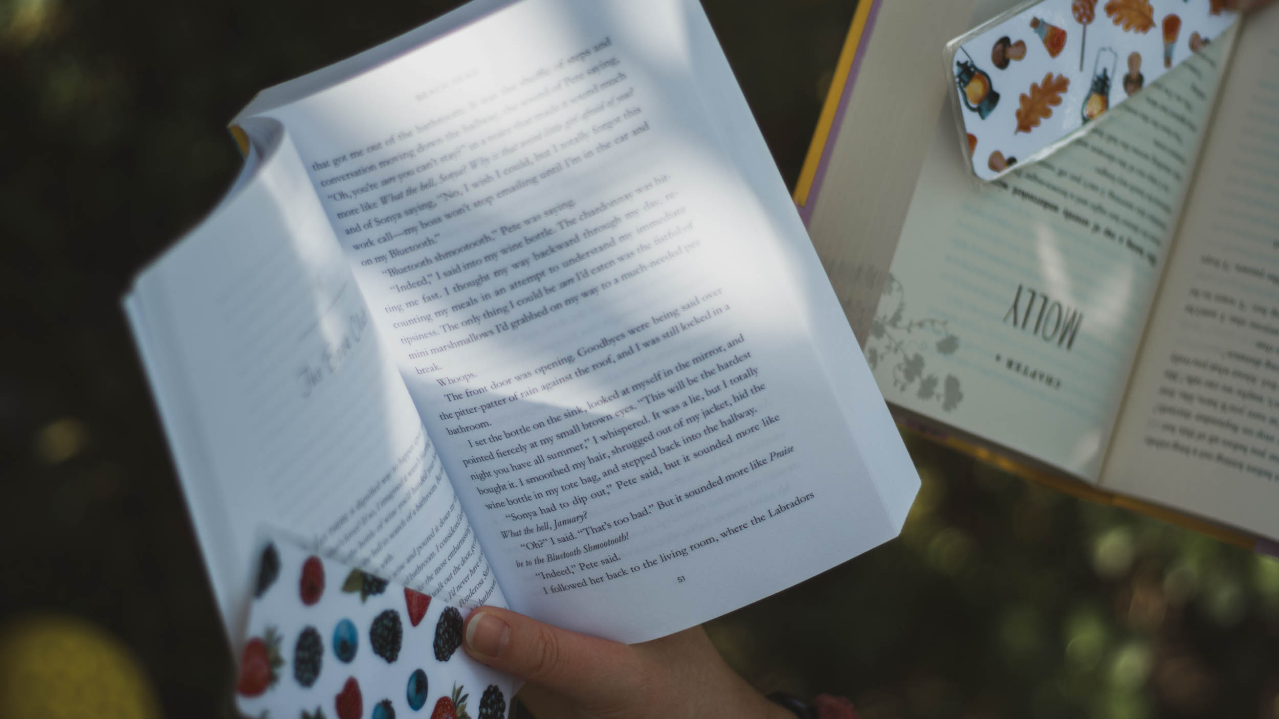 Photo of two open books being held up. One has a bookmark featuring different summer berries and the other has a bookmark featuring fall items like mushrooms, lanterns, and leaves.