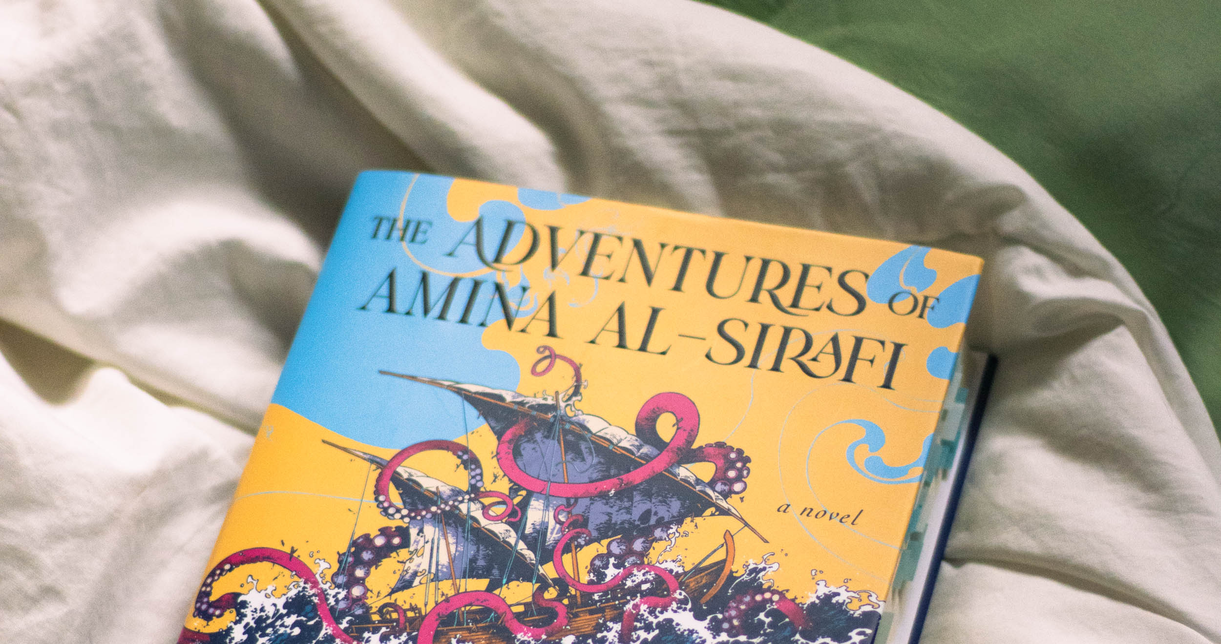 Photo of the book The Adventures of Amina Al-Sirafi up close on white and green fabric. The top of the book cover is visible and shows the title and a pirate ship surrounded by tentacles.