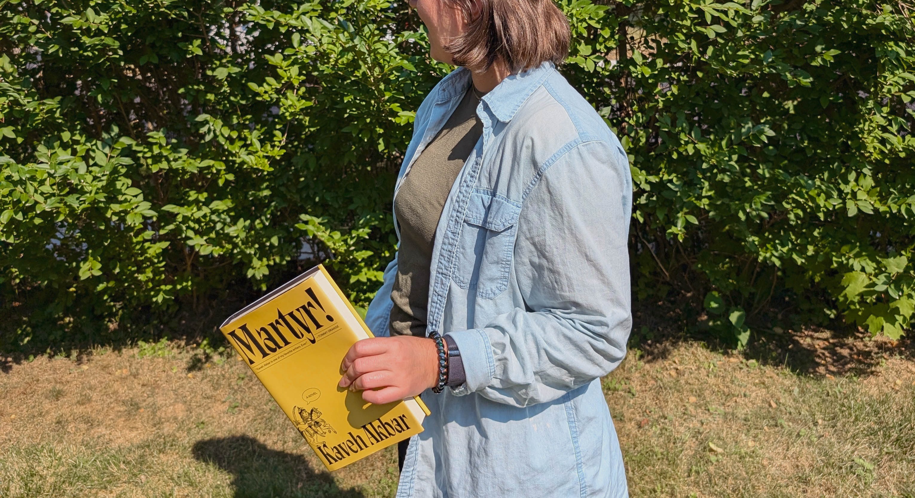 Photo of Bella standing in front of a leafy hedge looking away from the camera and holding the book Martyr! by Kaveh Akbar.