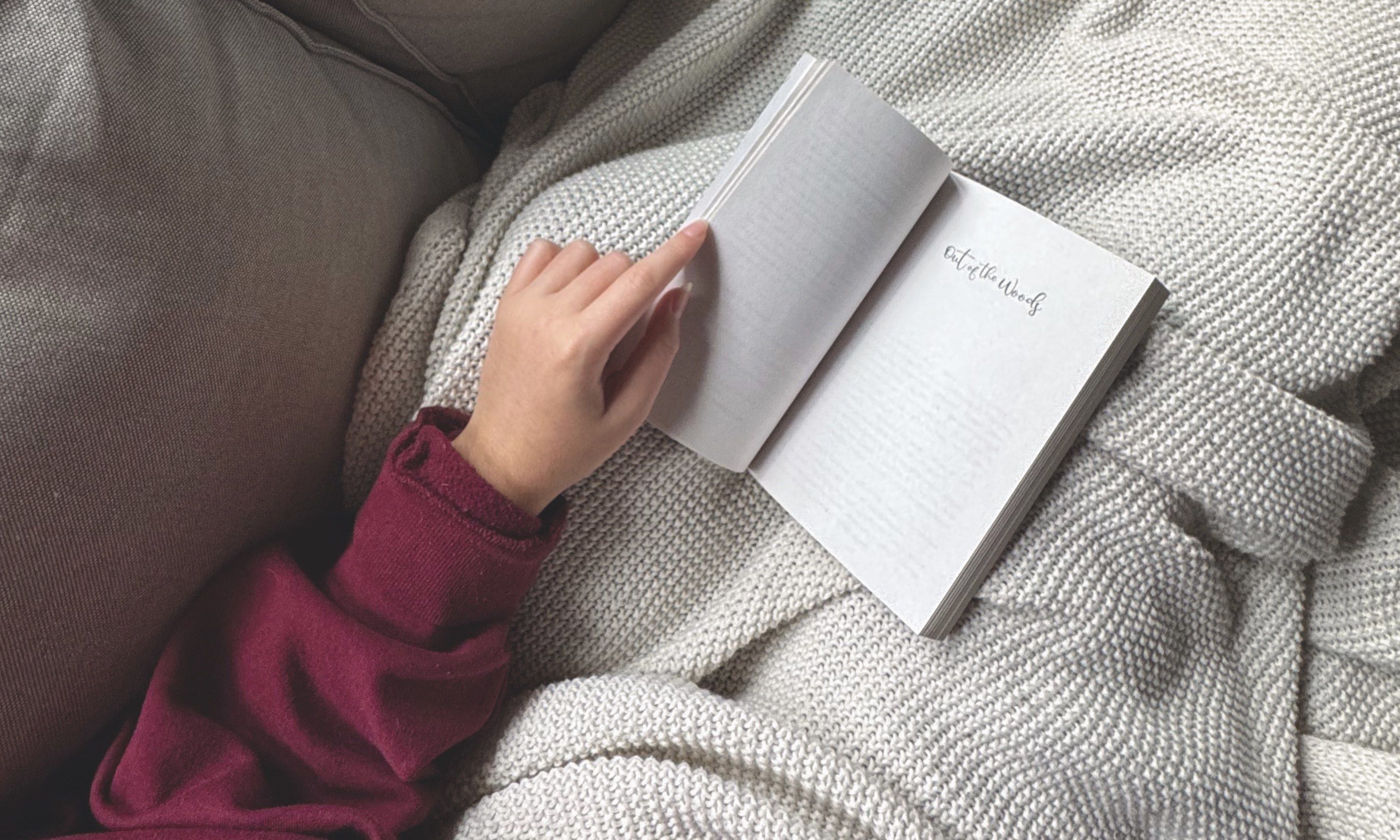 Photo of Bella's hand holding a book open to the title page which says Out of the Woods. The book is resting on Bella's lap which is covered in a white knitted blanket.